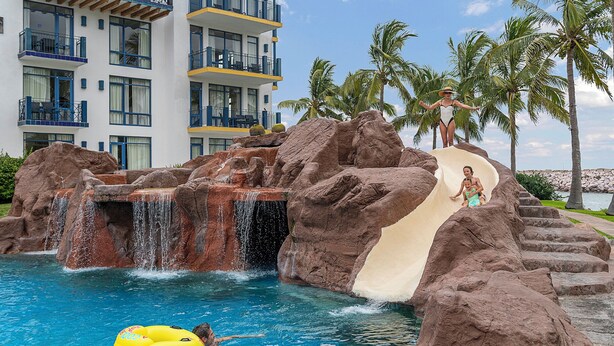 A pool area with a water slide, palm trees, and a building with balconies. - El Cid Marina Beach Hotel - All Inclusive (Mazatlán)