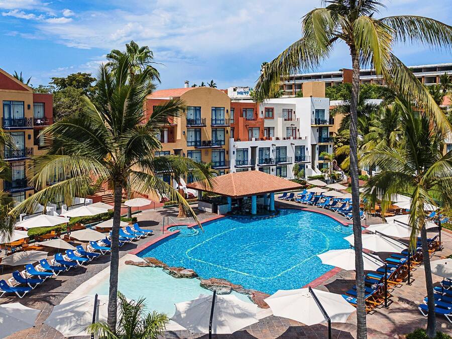 Un resort con una piscina centrale circondata da sdraio e ombrelloni, sullo sfondo palme e edifici a più piani.