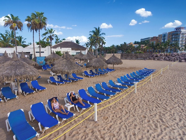 Une plage bordée de rangées de chaises longues bleues et d’ombrelles en chaume, avec des palmiers et un bâtiment en arrière-plan.