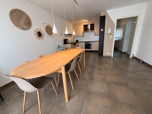 A modern kitchen with a dining area, featuring a long wooden table, chairs, pendant lights, and a visible oven. - Familytime (Oostende)