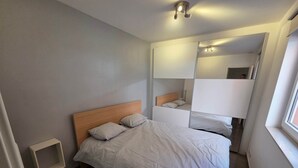 A bedroom with a bed, a bedside table, a mirror, and a window. - Familytime (Oostende)