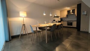 A modern kitchen with a dining area, featuring a table set for four, a standing lamp, and pendant lights. - Familytime (Oostende)