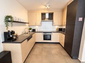 A modern kitchen with light wood cabinets, a stainless steel range hood, a built-in oven, a black refrigerator, and a coffee machine. - Family Time (Oostende)