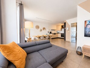 Una sala de estar moderna con un sofá gris, un comedor con mesa y sillas, una cocina con armarios de tonos claros, y un cartel en la pared.