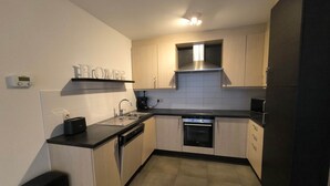 A compact kitchen with light wood cabinets, a black countertop, a built-in oven, and a stainless steel range hood. - Familytime (Oostende)