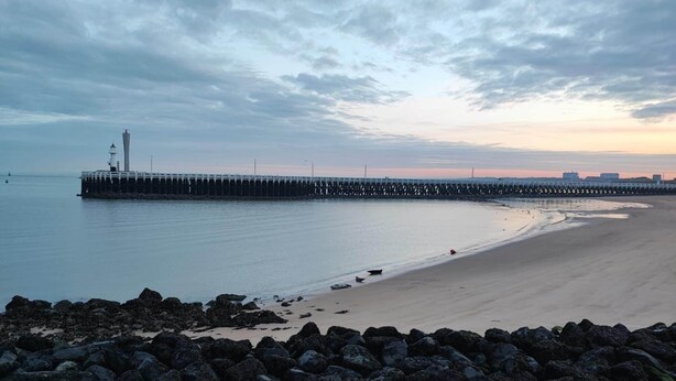 A coastal scene with a pier extending into calm waters, a sandy beach, and a rocky foreground. - Familytime (Oostende)
