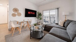 A modern living room with a grey sofa, a round coffee table, a dining area with a white table and chairs, a TV mounted on the wall, and a balcony with a view. - 4BR with Balcony and City View + Parking (Charlotte)