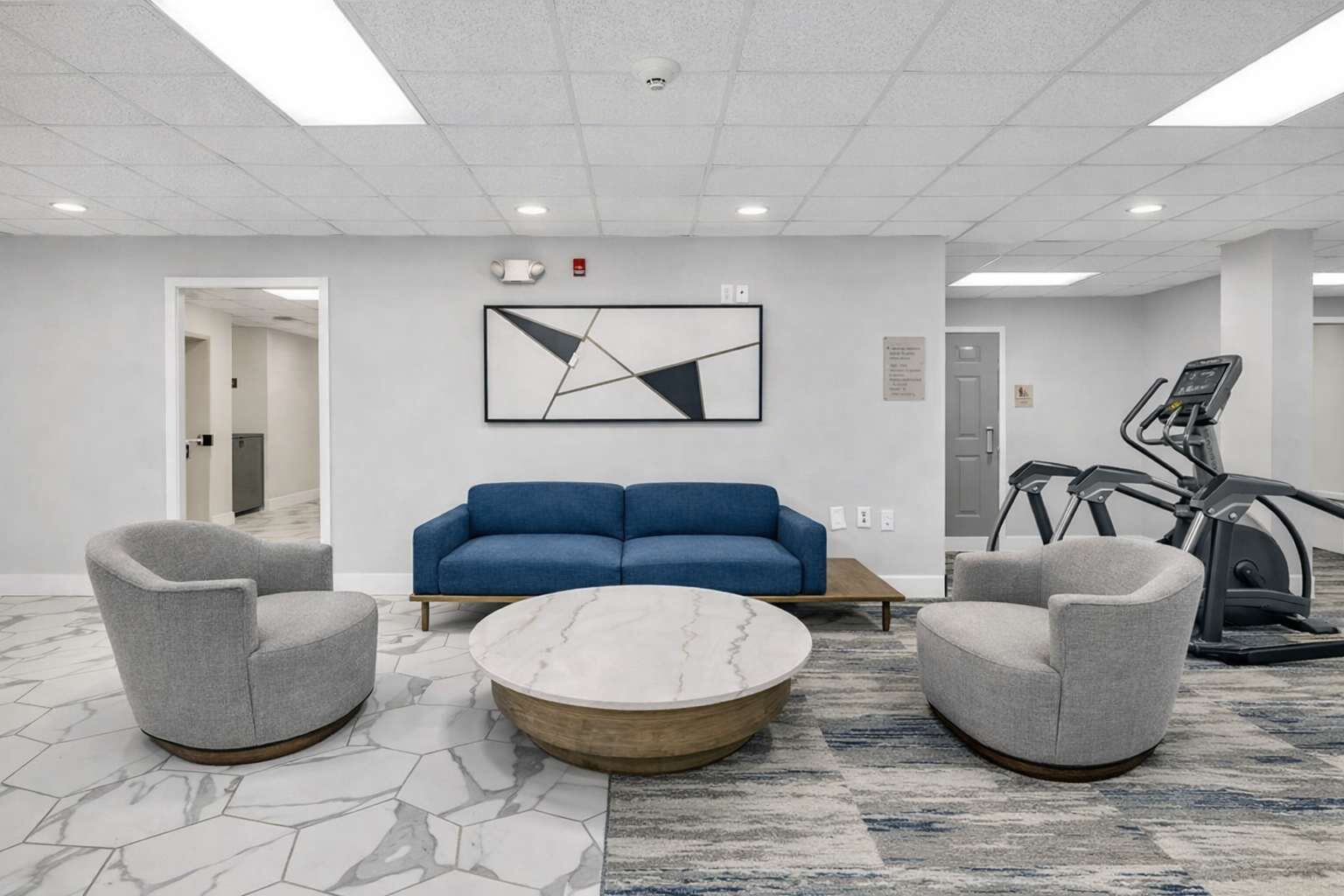 A modern lounge area with a blue sofa, two grey armchairs, a marble coffee table, and a treadmill.