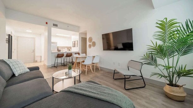 A modern living room with a sofa, coffee table, and a TV mounted on the wall. There is a dining area with chairs and a table, and a kitchen in the background. - Lux 3BR w 3 King Beds, Downtown, Parking (Charlotte)