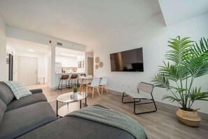 A modern living room with a sofa, coffee table, and a TV mounted on the wall. There is a dining area with chairs and a table, and a kitchen in the background. - Lux 3BR w 3 King Beds, Downtown, Parking (Charlotte)