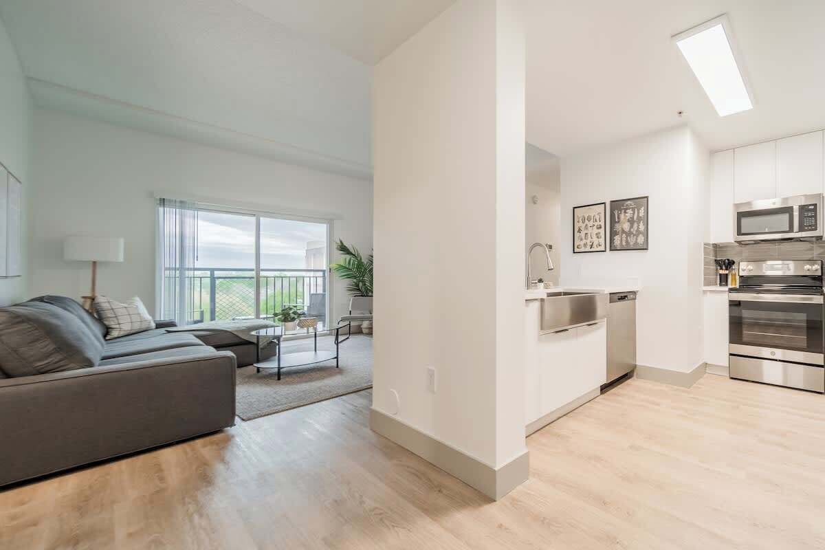 Un appartement moderne avec un canapé gris, une petite table à manger, une cuisine équipée d’appareils électroménagers en acier inoxydable et de grandes fenêtres offrant une vue dégagée.