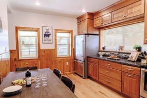 Une cuisine avec des meubles de rangement en bois, un réfrigérateur en acier inoxydable, une table à manger avec des chaises et une fenêtre avec des stores.