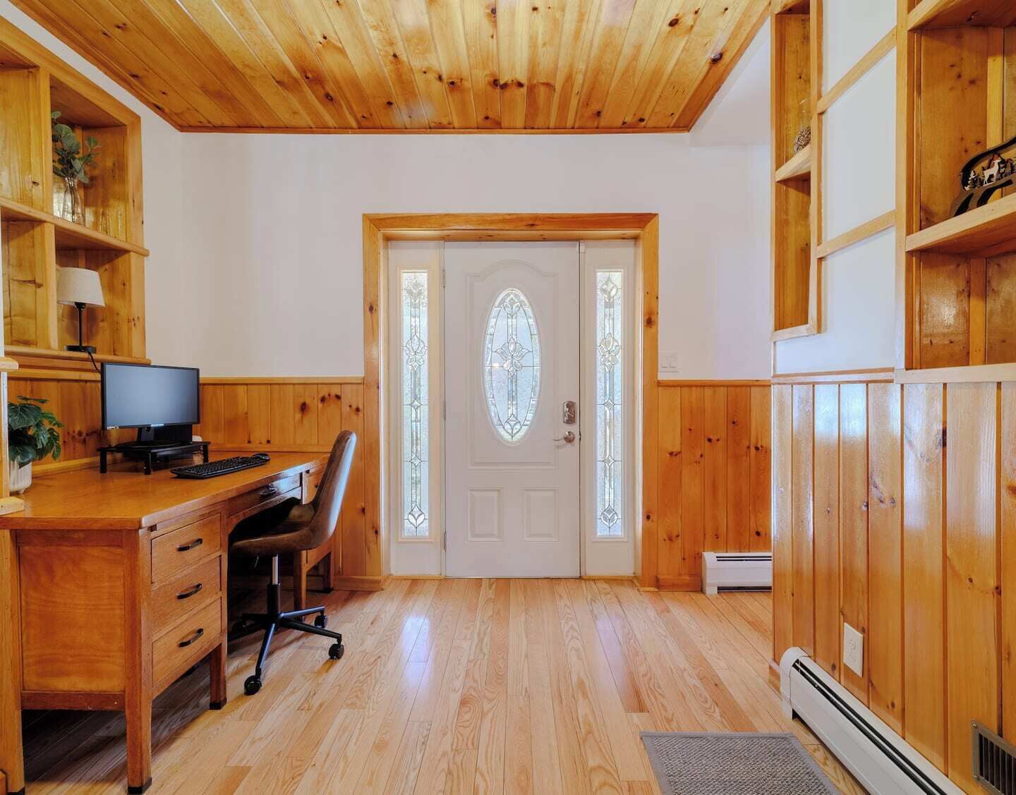 A room with wooden walls and ceiling, a desk with a computer, a chair, and a door with a glass panel.