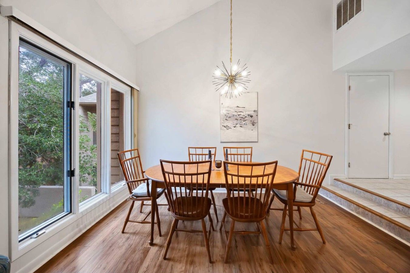 A dining area with a round table and six chairs, large windows, and a hanging light fixture.