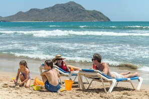 A beach scene with people relaxing on sun loungers, a child playing with sand toys, and a mountain in the background. - El Cid El Moro Beach Hotel - All Inclusive (Mazatlán)
