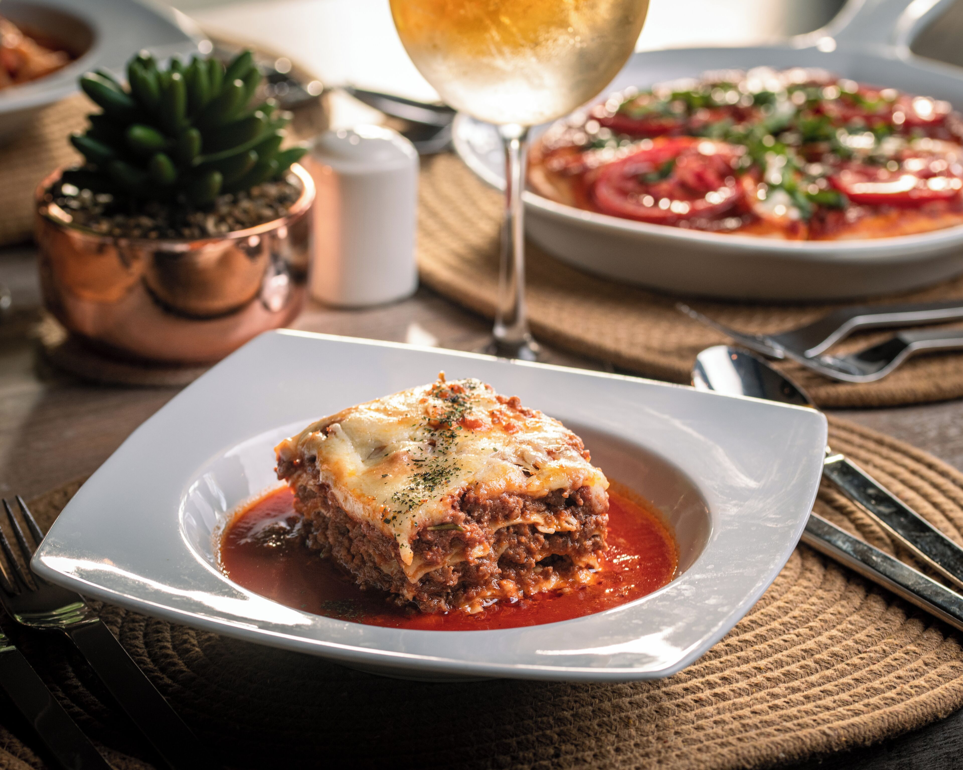 A plate of lasagna with a side of tomato salad, a glass of wine, and a small potted plant on a table.