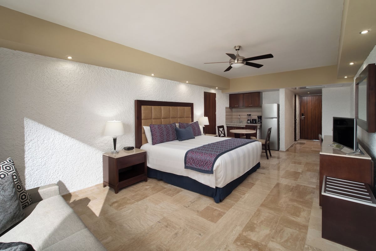 A hotel room with a bed, bedside table, lamp, and a kitchenette area.