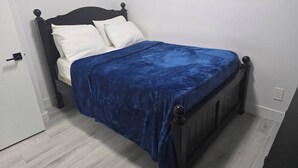 A bed with a dark headboard, white pillows, and a blue bedspread in a room with light wooden flooring and white walls. - Cy Becker Drive 2023 Luxury Home (Edmonton)