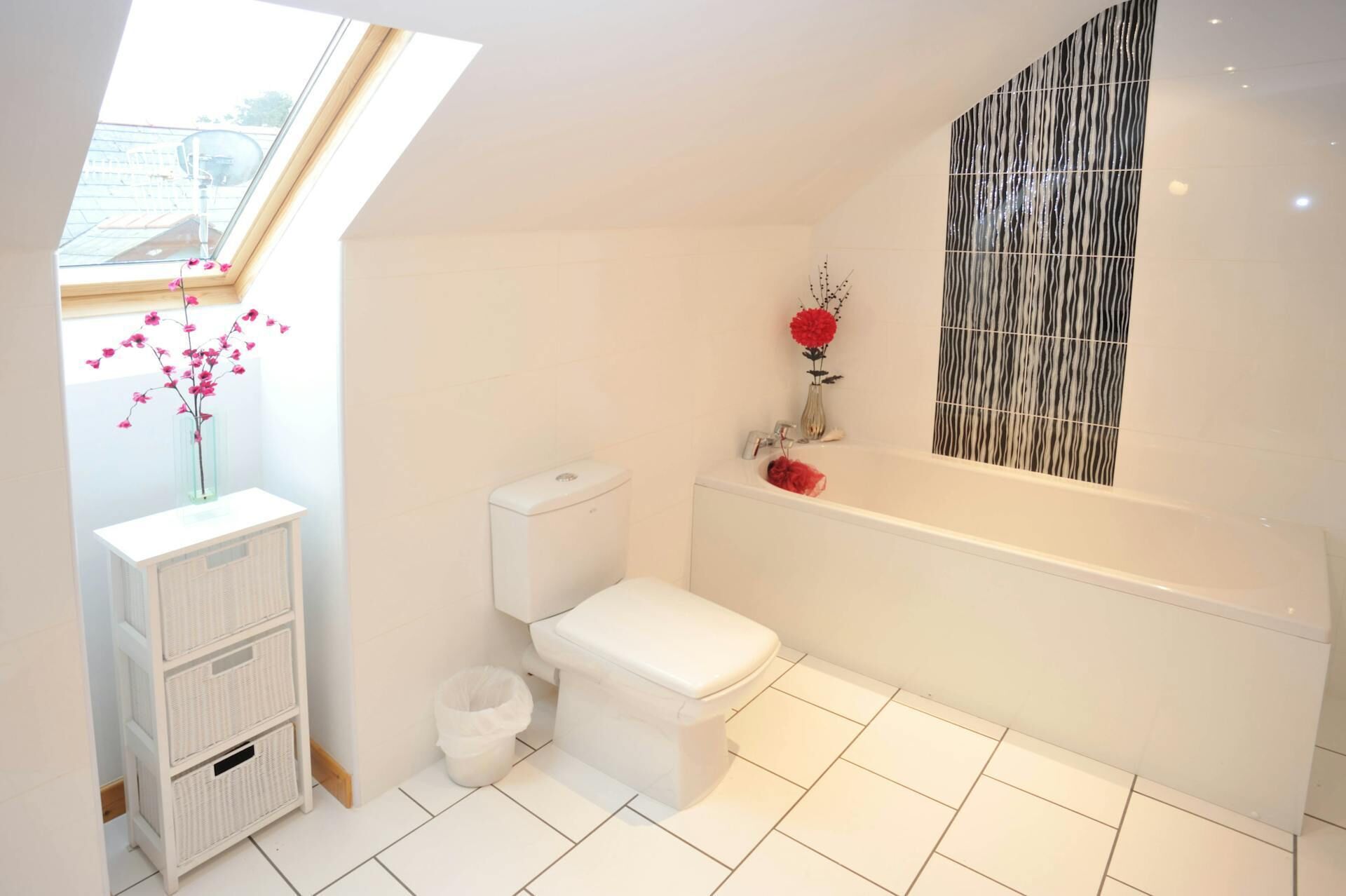 A modern bathroom with a skylight, white tiles, a toilet, a bathtub, and a small trash bin.
