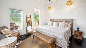 A spacious bedroom with a large bed, a wooden bench, a wicker chair, and a view of the outdoors through a large window. - Saddle Hill View Villa (Charlestown)