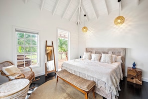 A spacious bedroom with a large bed, a wooden bench, a wicker chair, and a view of the outdoors through a large window. - Saddle Hill View Villa (Charlestown)