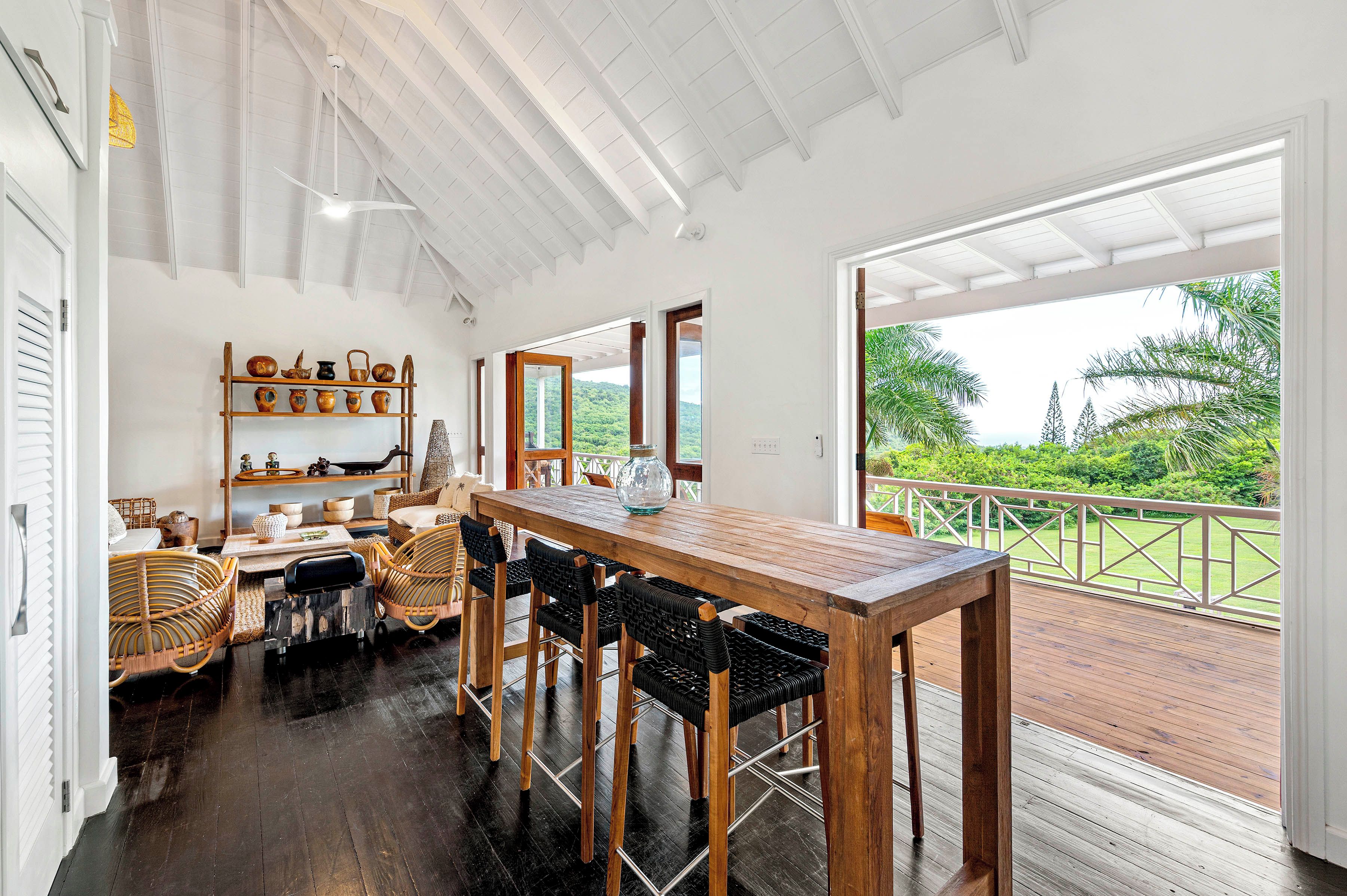 A spacious dining area with a wooden table and chairs, a shelf with pottery, and a view of lush greenery through large windows.