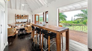 A spacious dining area with a wooden table and chairs, a shelf with decorative items, and a view of lush greenery through large windows. - Saddle Hill View Villa (Charlestown)