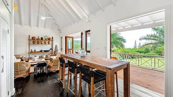 A spacious dining area with a wooden table and chairs, a shelf with pottery, and a view of lush greenery through large windows.