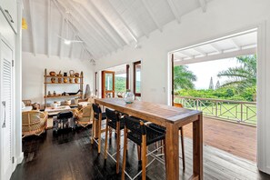 A spacious dining area with a wooden table and chairs, a shelf with decorative items, and a view of lush greenery through large windows. - Saddle Hill View Villa (Charlestown)
