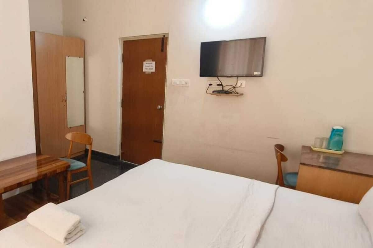 A hotel room with a bed, a wooden desk, a chair, a television mounted on the wall, and a small table with toiletries.
