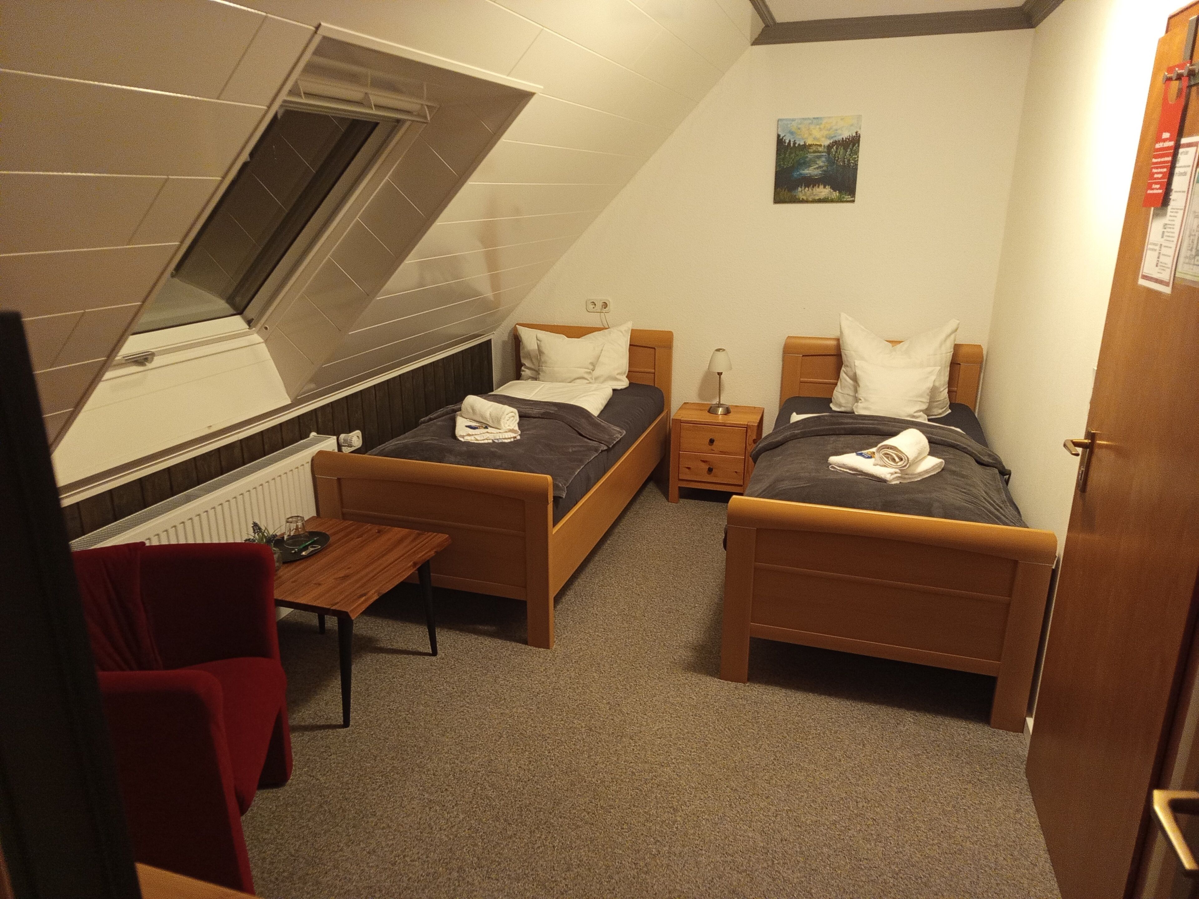 A small, well-lit room with two single beds, a small table, and a red chair.
