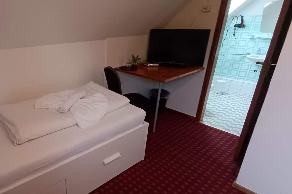 Un lit simple avec une literie blanche, un bureau avec un ordinateur et une plante, et un sol recouvert d’un tapis rouge.
