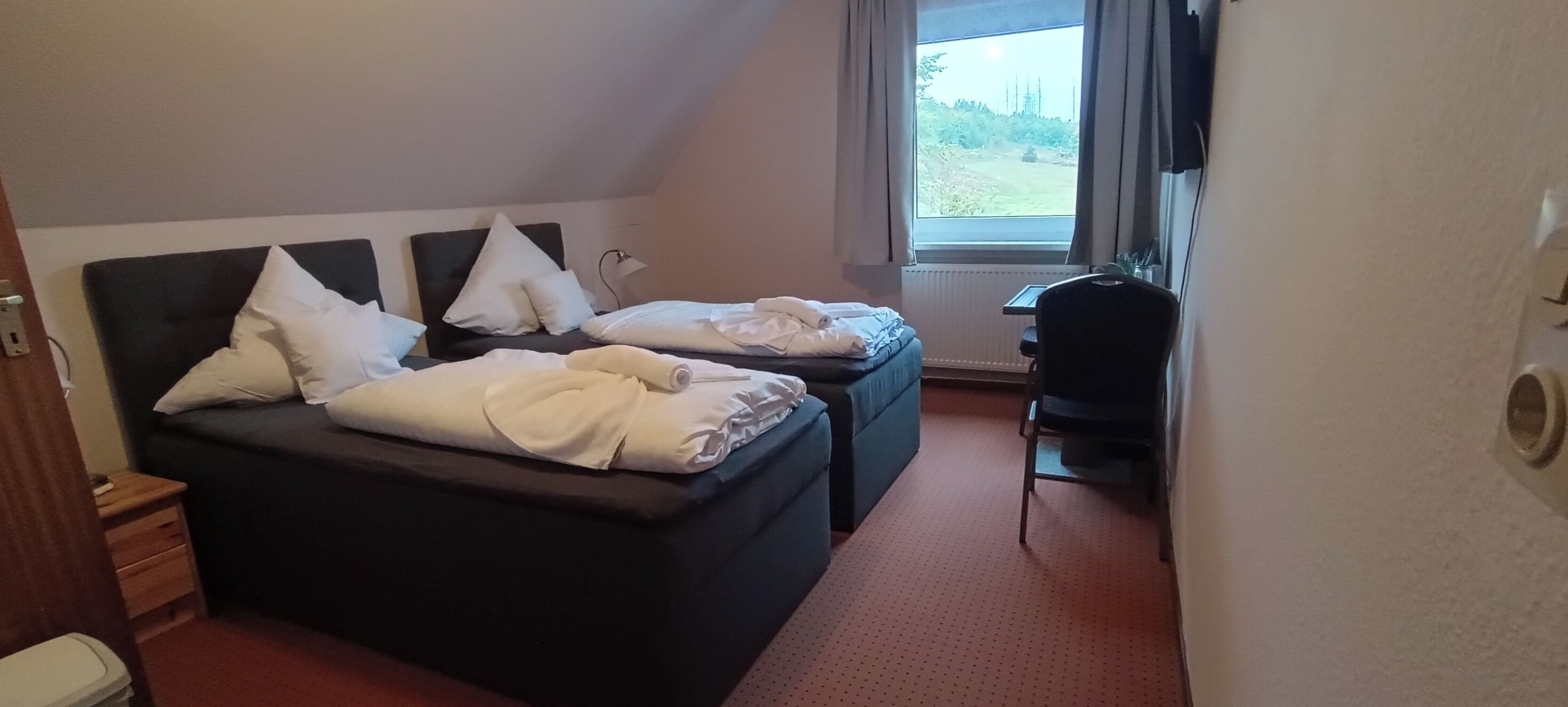 Ein Hotelzimmer mit zwei Betten, einem Schreibtisch und einem Fenster mit Blick auf Grünflächen.