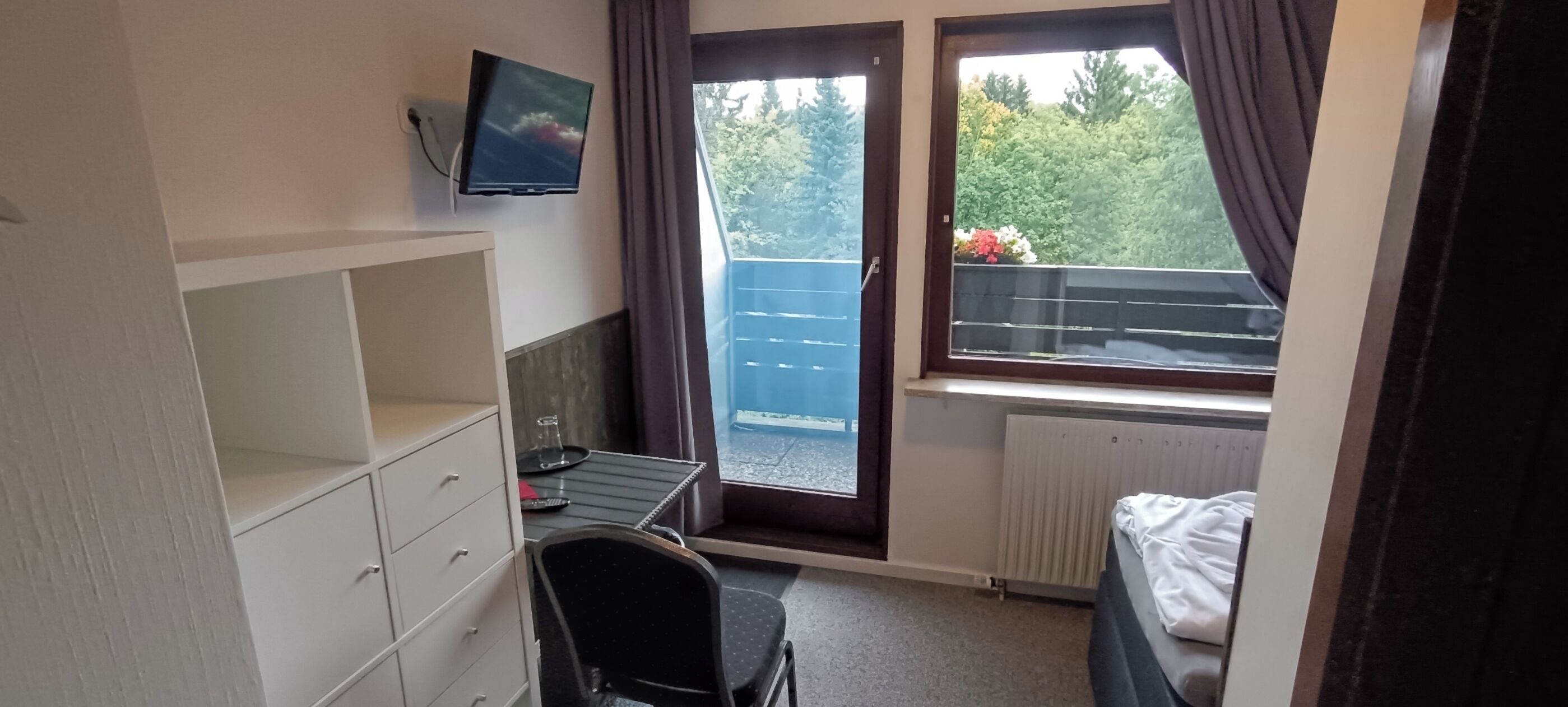 A small room with a desk, chair, and a TV mounted on the wall. There is a window with a view of greenery outside.