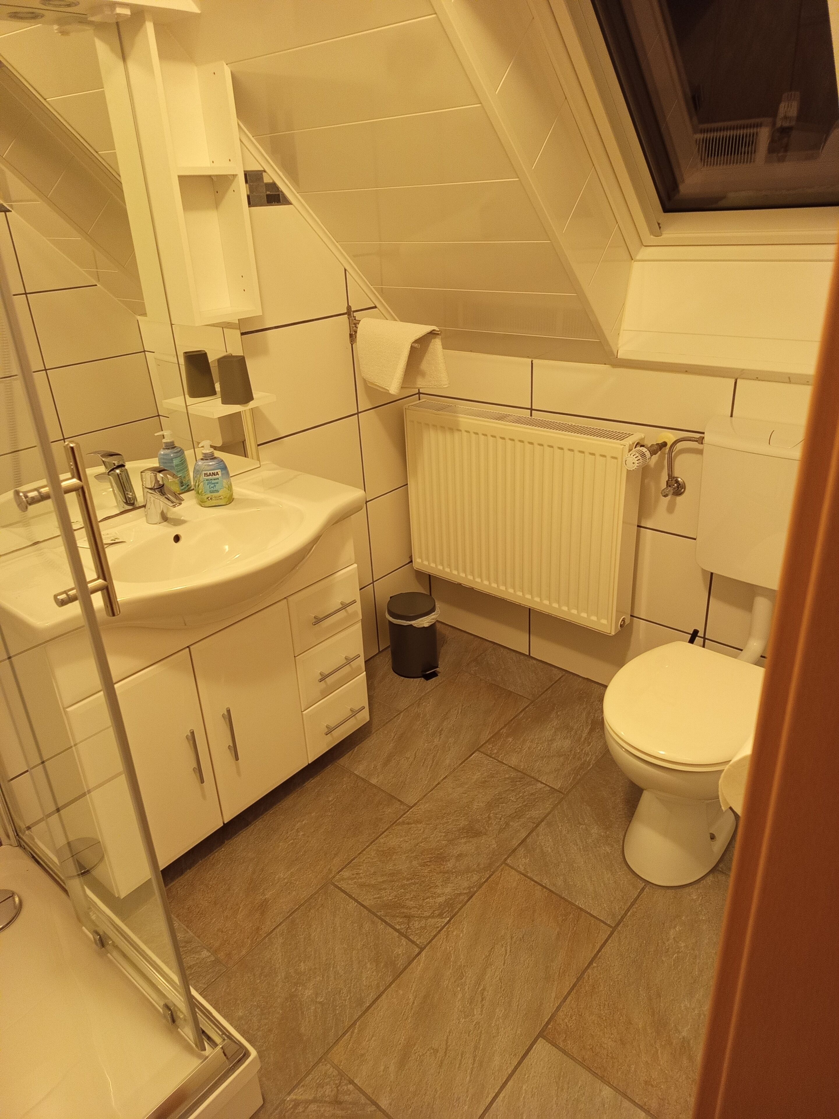 Un baño moderno con lavamanos, espejo y retrete blancos. Hay un radiador sobre el retrete y un pequeño cubo de basura a su lado.