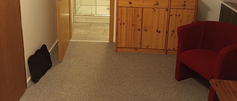 Een knus, klein kamertje met een douche, een televisie op een houten kast en een rode stoel.