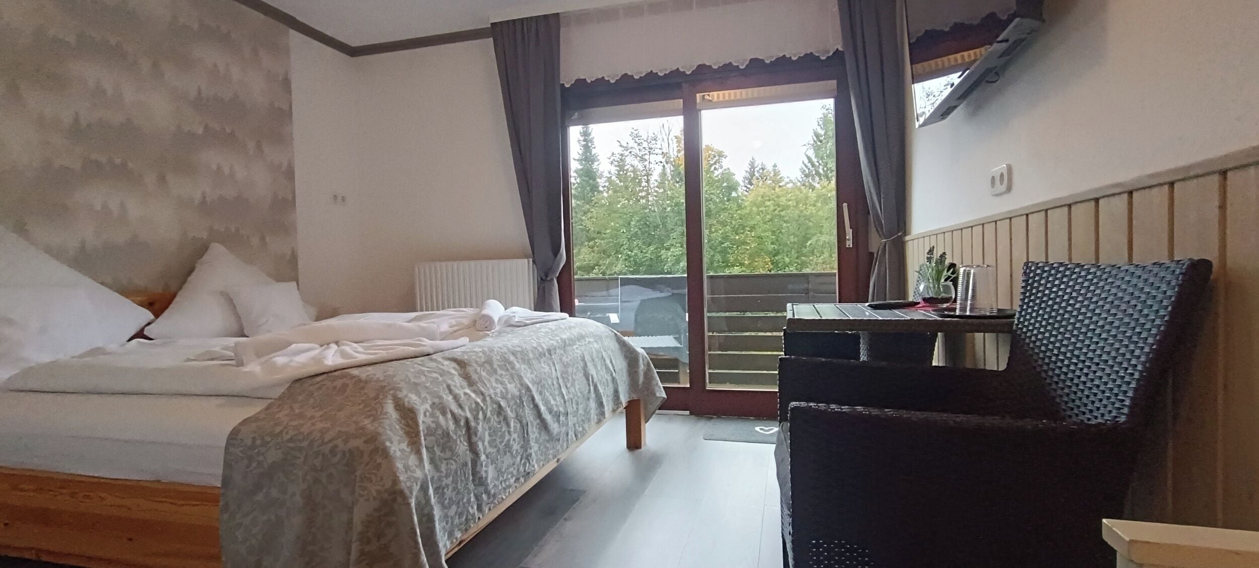 A bedroom with a bed, a desk, and a chair. There is a window with curtains and a view of trees outside.
