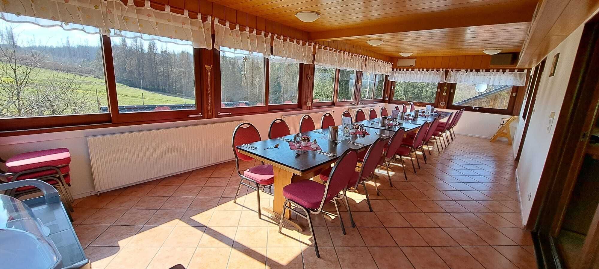 Un espace repas avec une longue table et des chaises, de grandes fenêtres avec des rideaux et une vue sur un paysage verdoyant.