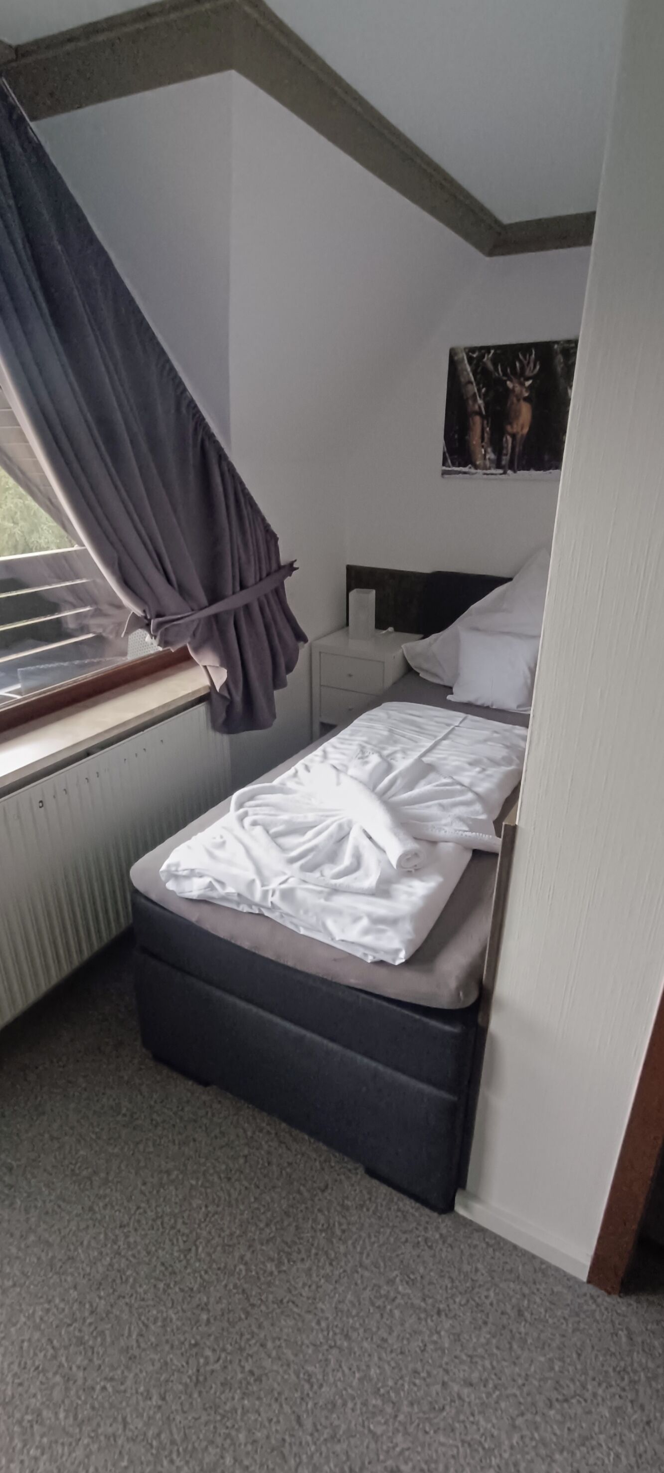 Una habitación pequeña con cama individual, ventana, radiador, cabecero y un cuadro colgado en la pared.