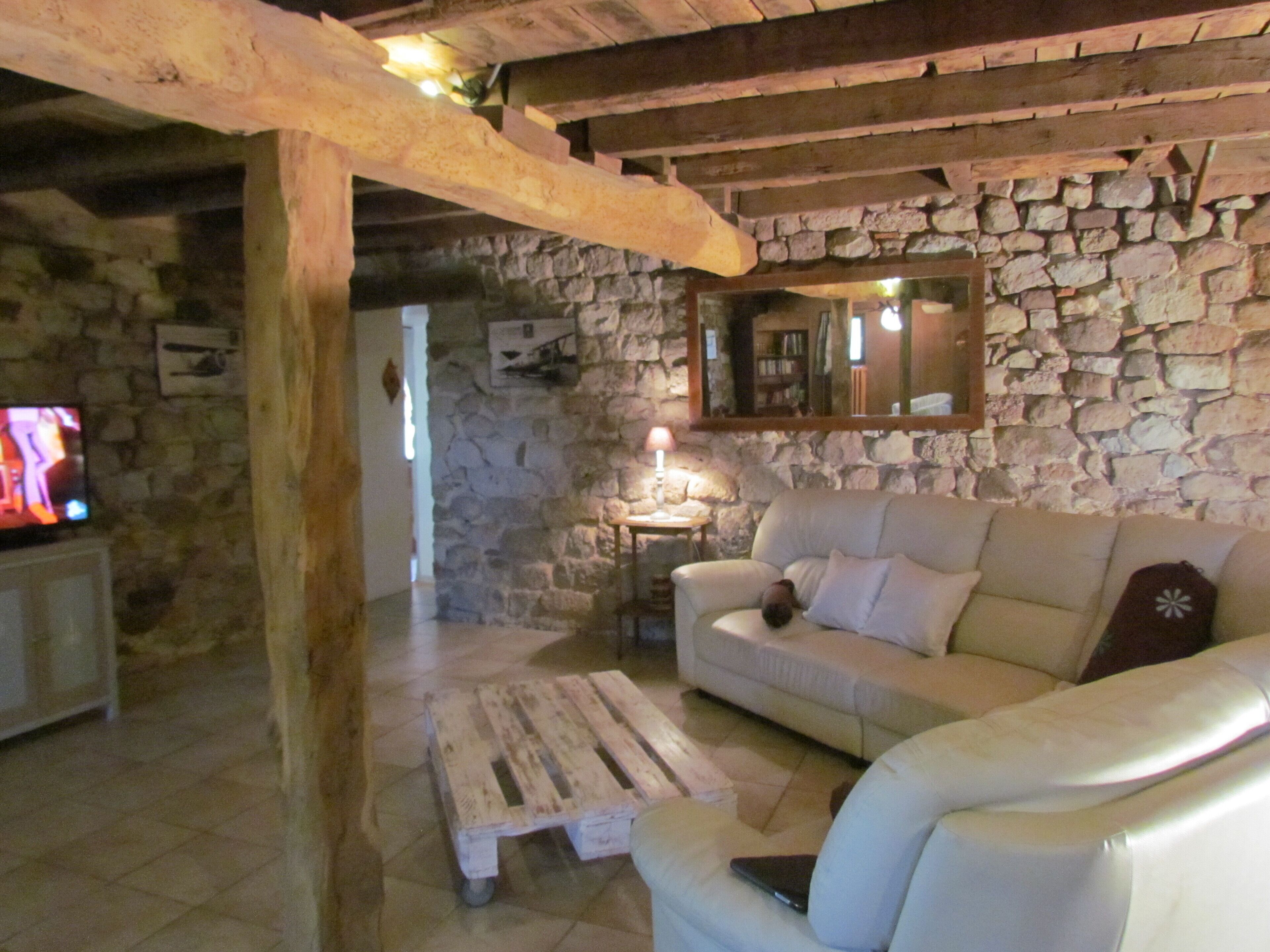 Una sala rústica con pared de piedra, techo de madera, un sofá blanco, una mesa de centro de madera y un televisor sobre un mueble.