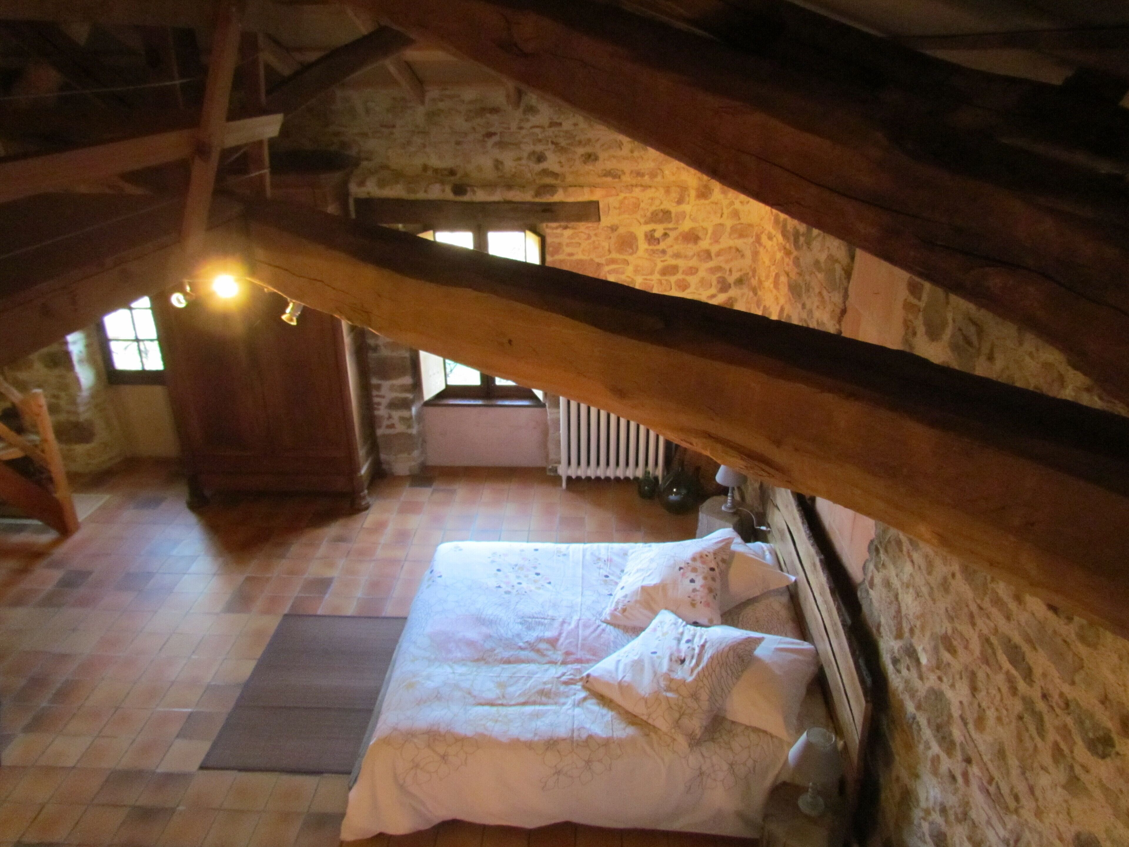 Een gezellig slaapkamer op zolder met een stenen muur, houten balken, een bed met wit beddengoed en uitzicht naar buiten door een raam.