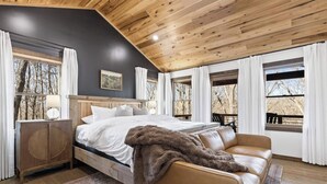 Ein Schlafzimmer mit Bett, einem Holz-Kopfbrett, einem braunen Ledersofa, einem Rattan-Nachttisch und einem großen Fenster mit Vorhängen.
