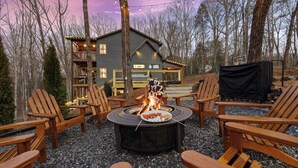 Ein gemütlicher Sitzbereich im Freien mit Feuerstelle, umgeben von Adirondack-Stühlen, vor einer rustikalen Holzhütte mit einer Veranda und Lichterketten.