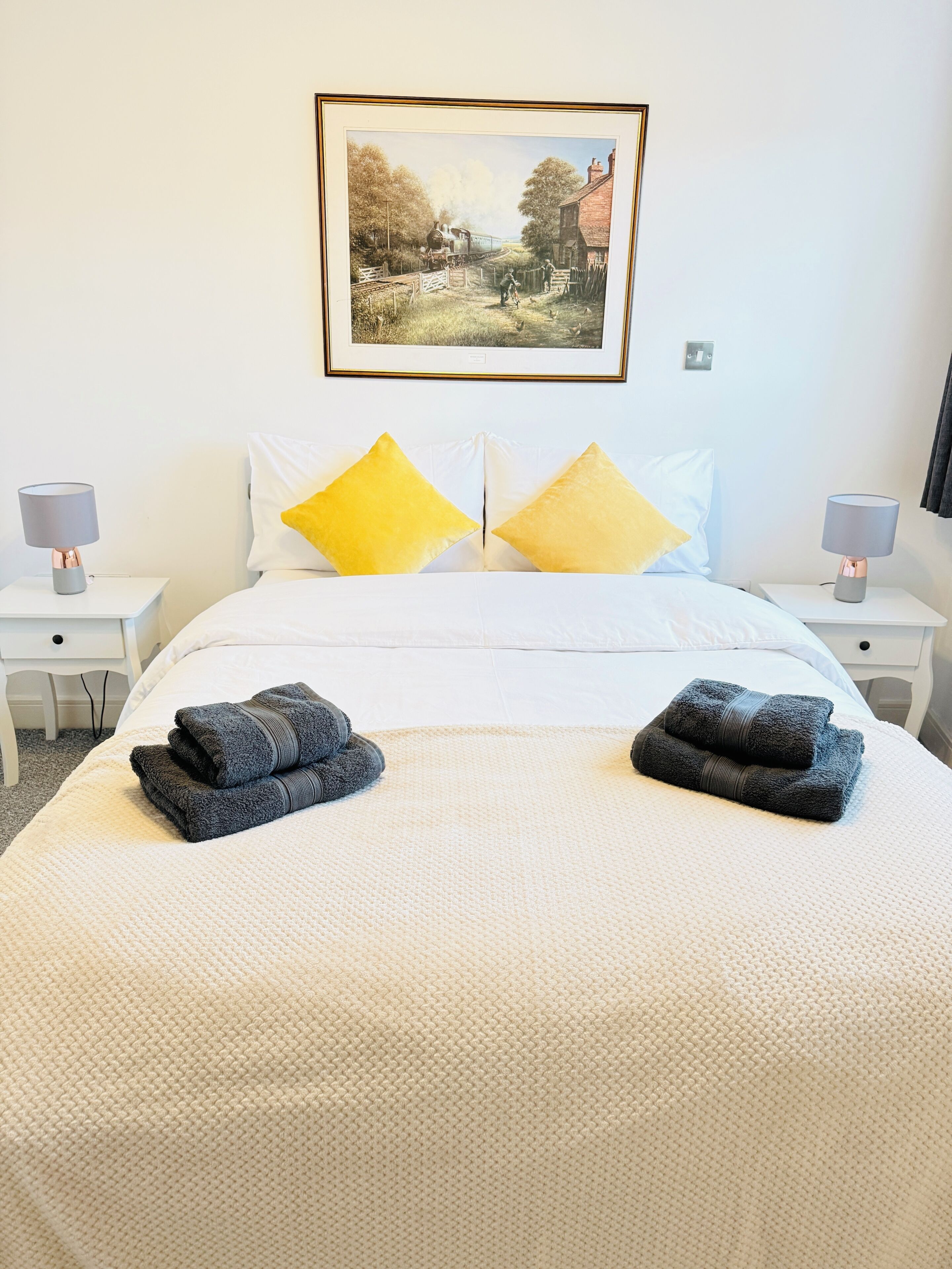 A hotel room with a bed, two nightstands, a lamp on each, a framed picture on the wall, and two towels on the bed.