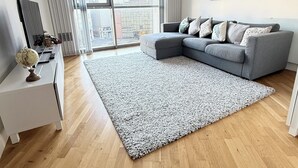 A modern living room with a grey sectional sofa, a white side table, a large grey rug, and a cityscape painting on the wall. - Modern 1-Bed Flat | Cardiff City Centre | Free Parking (Cardiff)