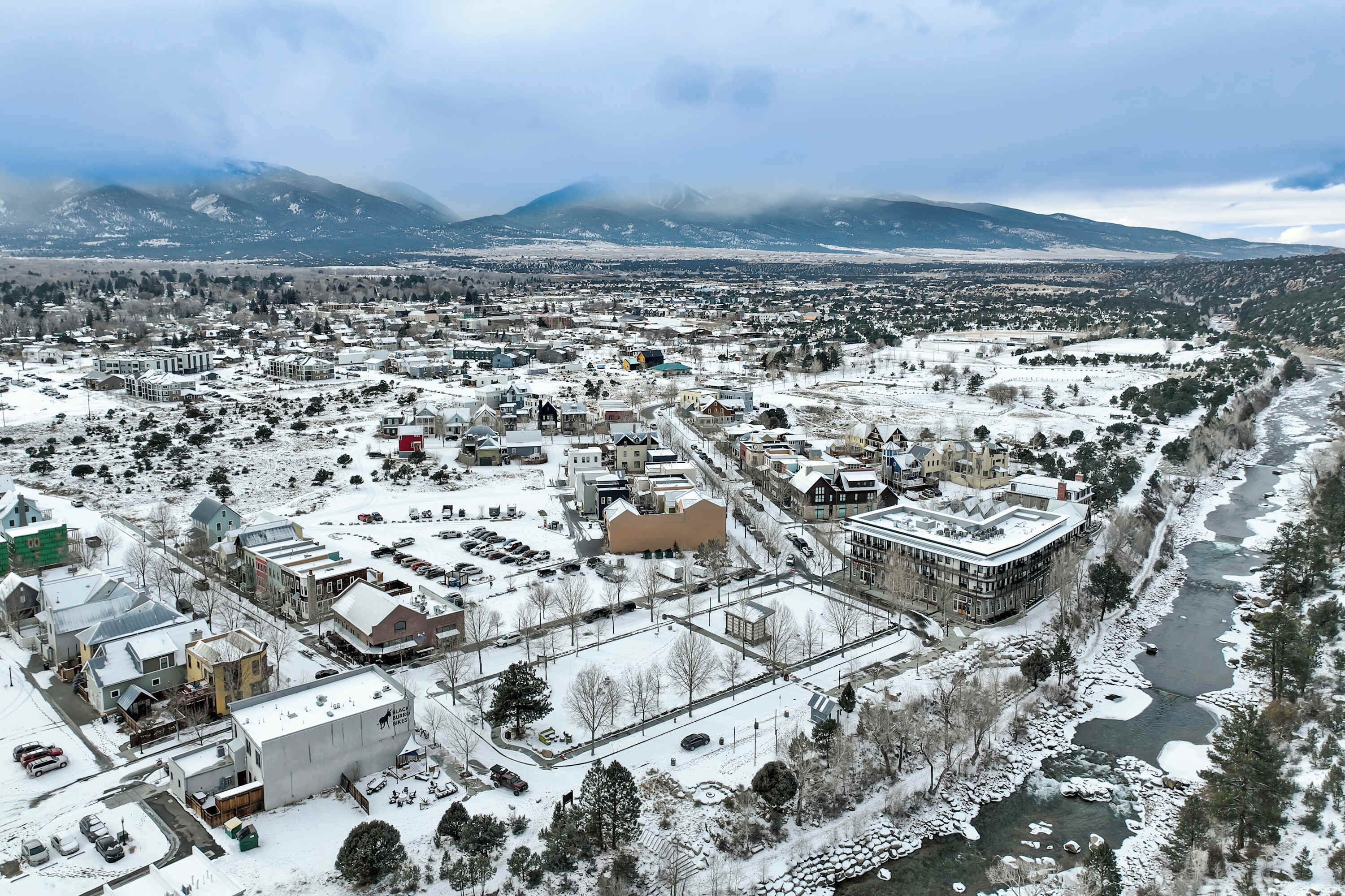 Un pueblo cubierto de nieve con un río, edificios y montañas al fondo.