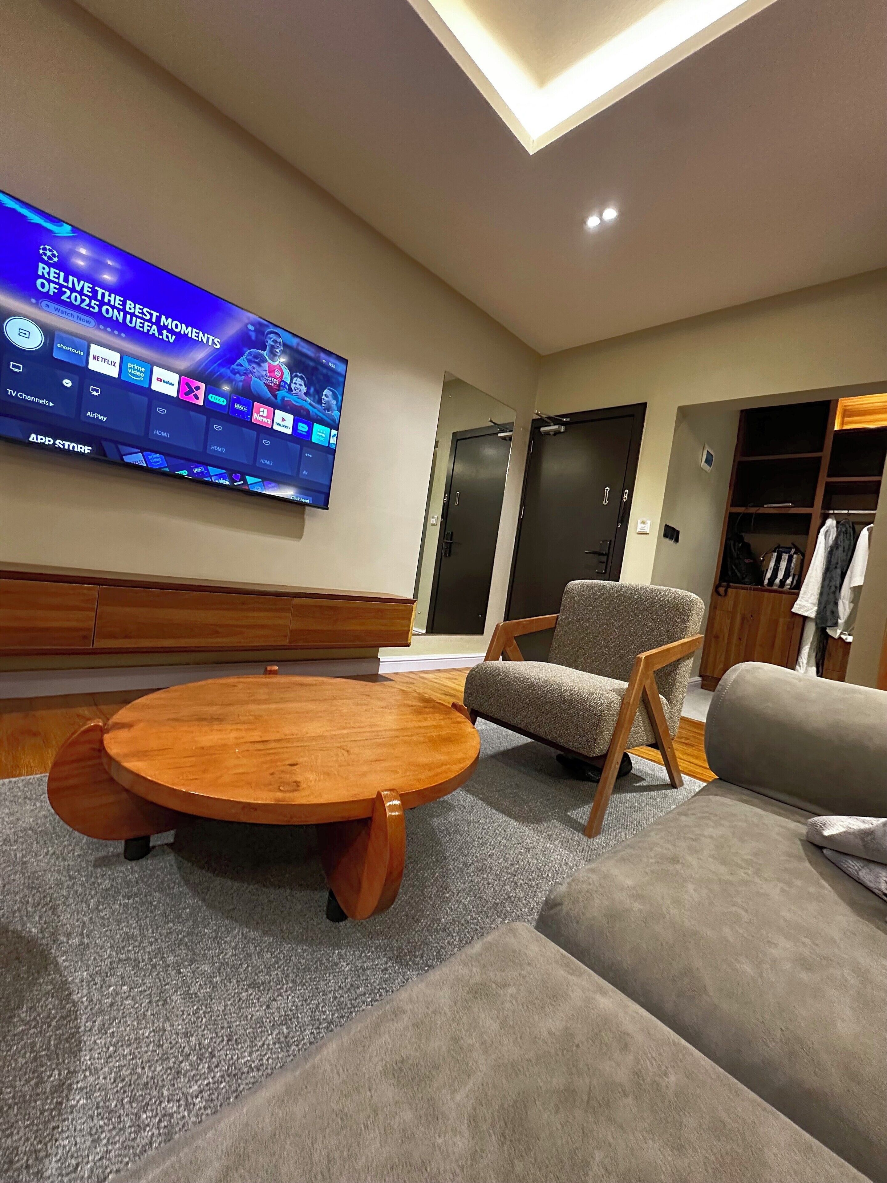 Ett modernt vardagsrum med en platt-TV som visar UEFA.tv, ett soffbord i trä, en grå fåtölj och en grå soffa.