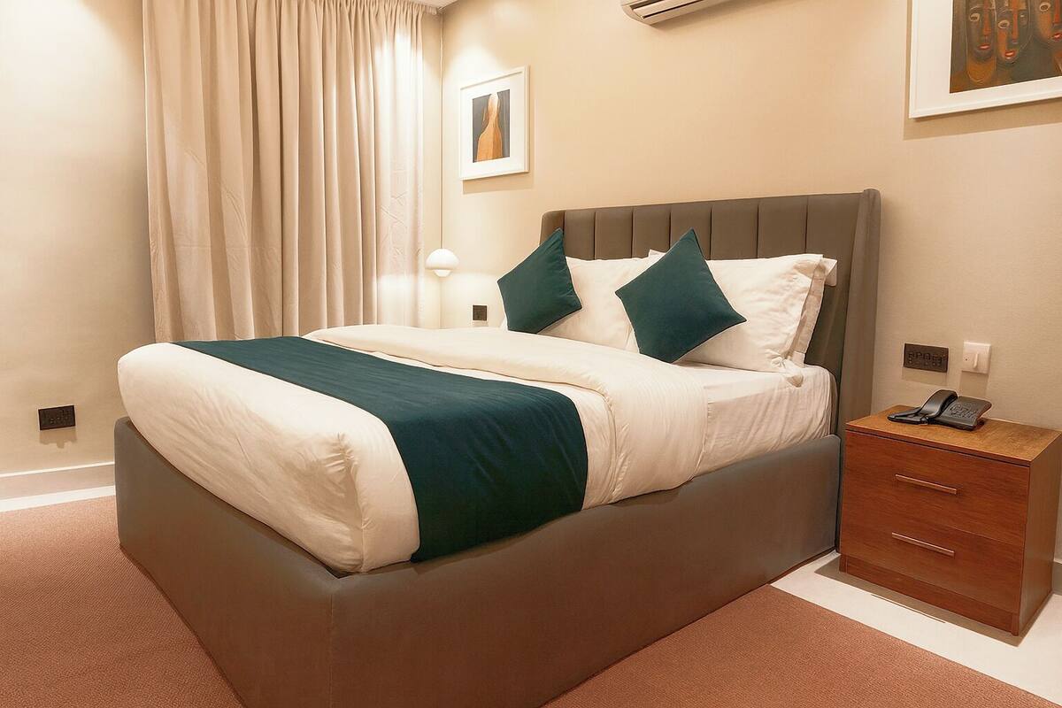 A neatly made bed with a brown headboard, a bedside table with a phone, and a wall-mounted air conditioner.