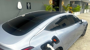 Hopeanvärinen sähköauto latautuu modernin rakennuksen edessä, ja kyltissä kerrotaan, että sähköautojen lataus on mahdollista.