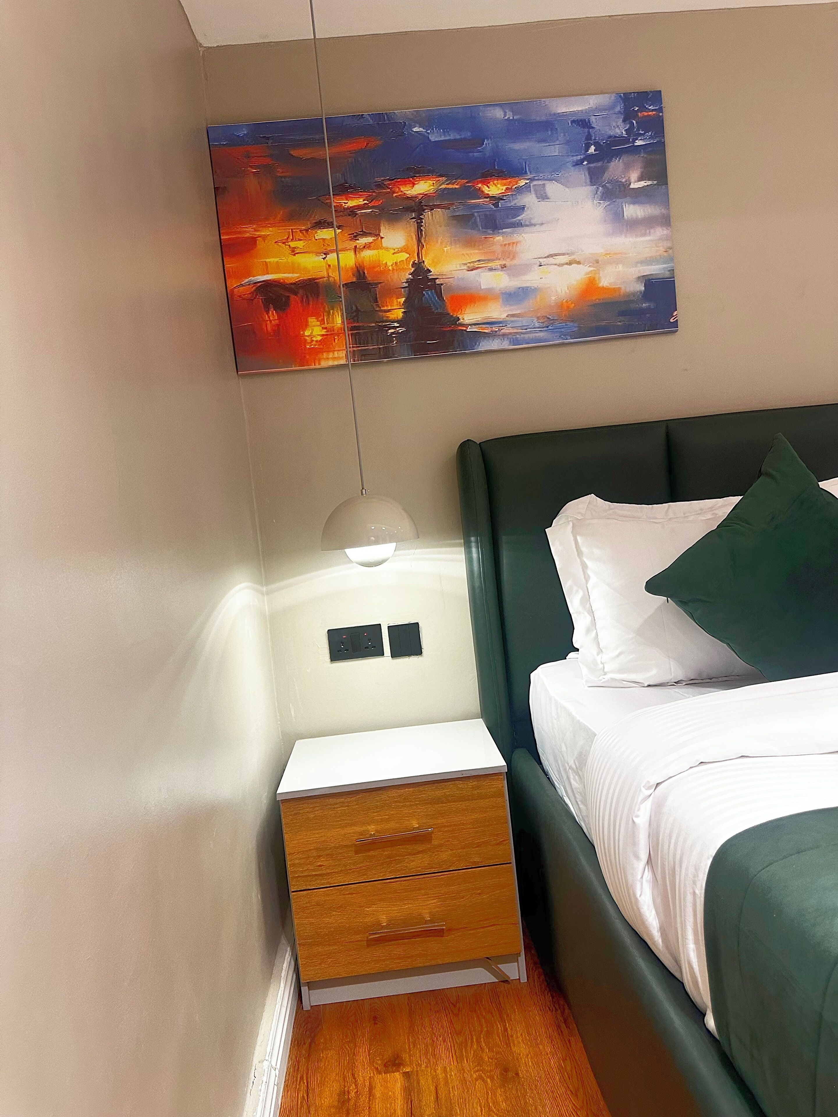 A hotel room with a bed, a nightstand with a lamp, a drawer, and a painting of the Eiffel Tower.